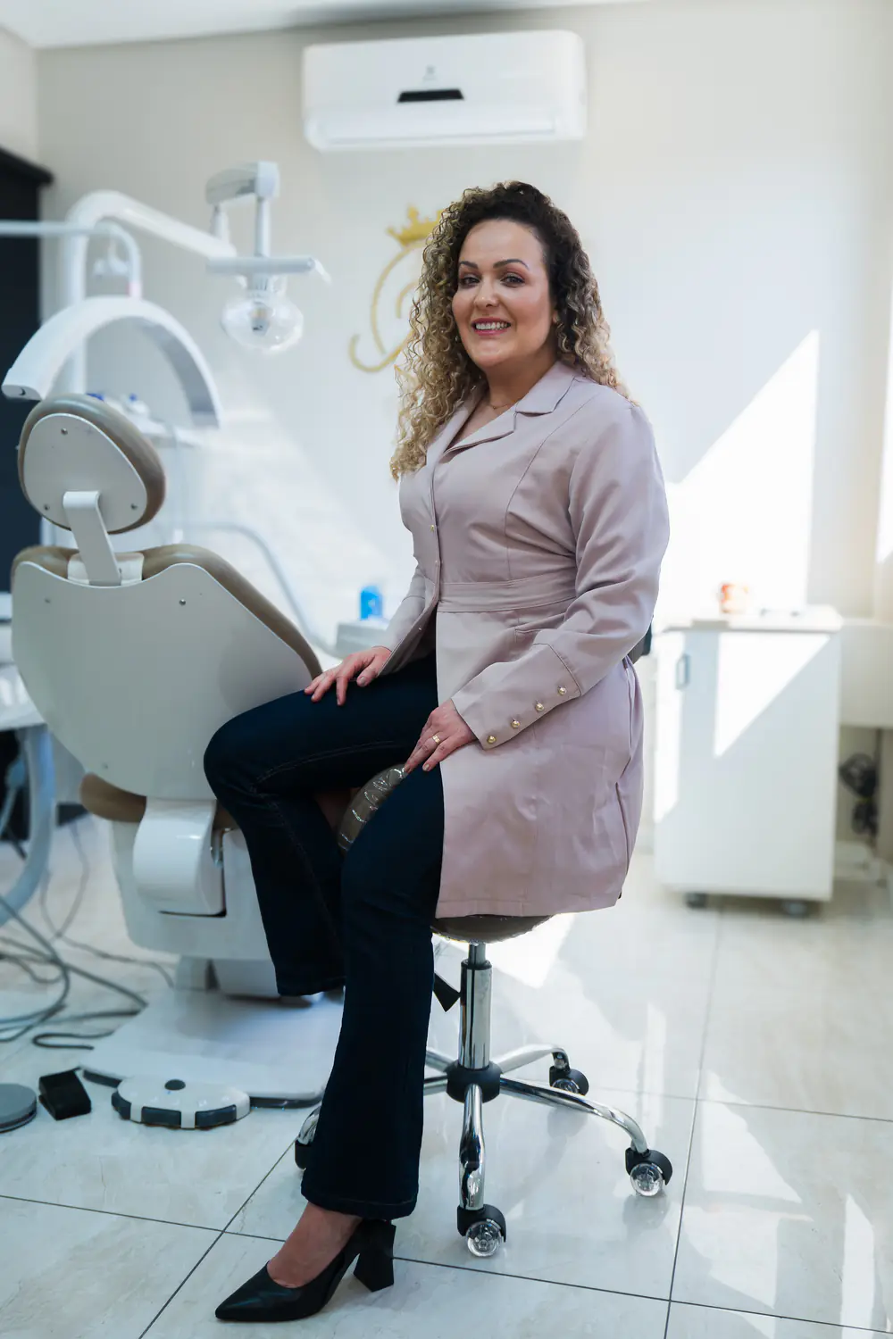 Dra. Jessica Koch sentada ao lado da cadeira odontológica na sala de atendimento, iluminada por luz natural